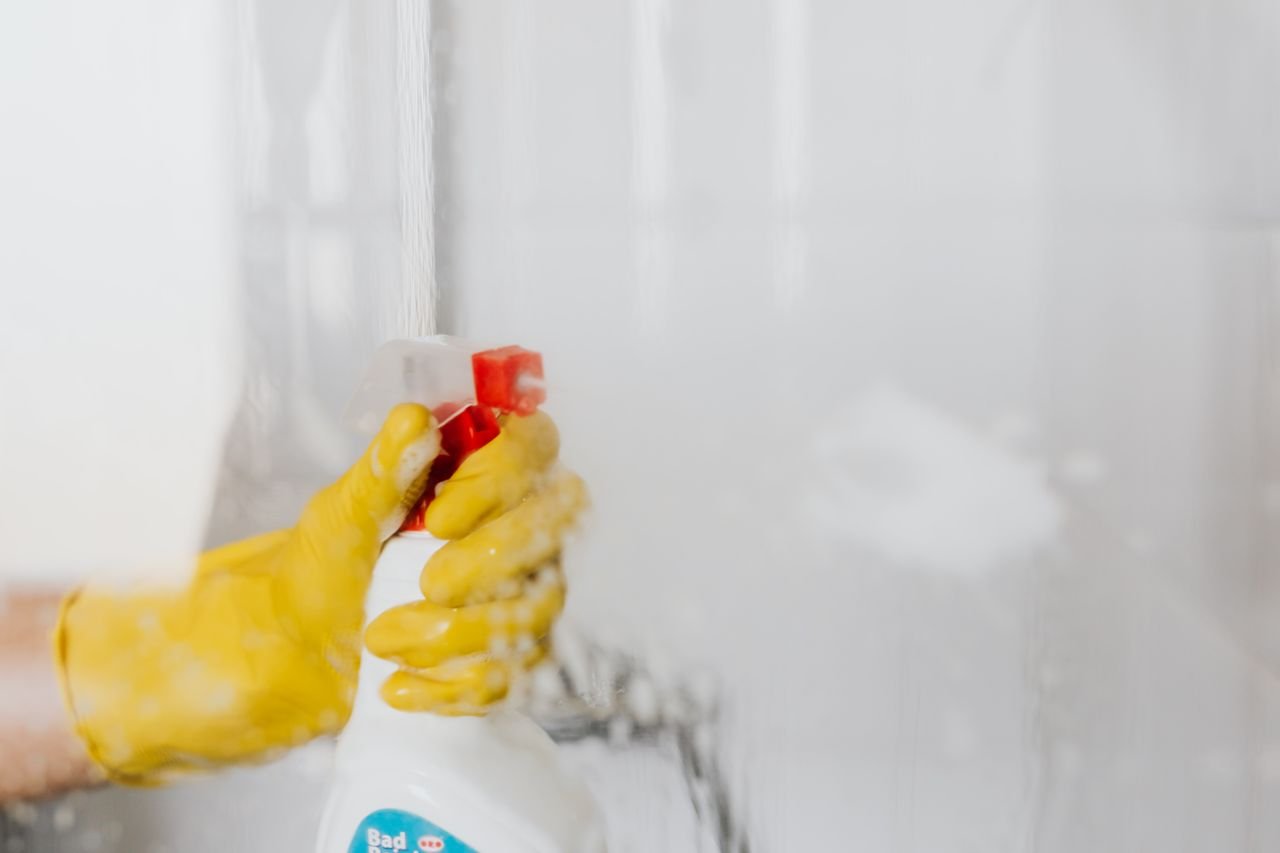 Crop unrecognizable person in yellow latex protective glove spraying detergent from bottle with plastic dispenser at glass of shower cabin